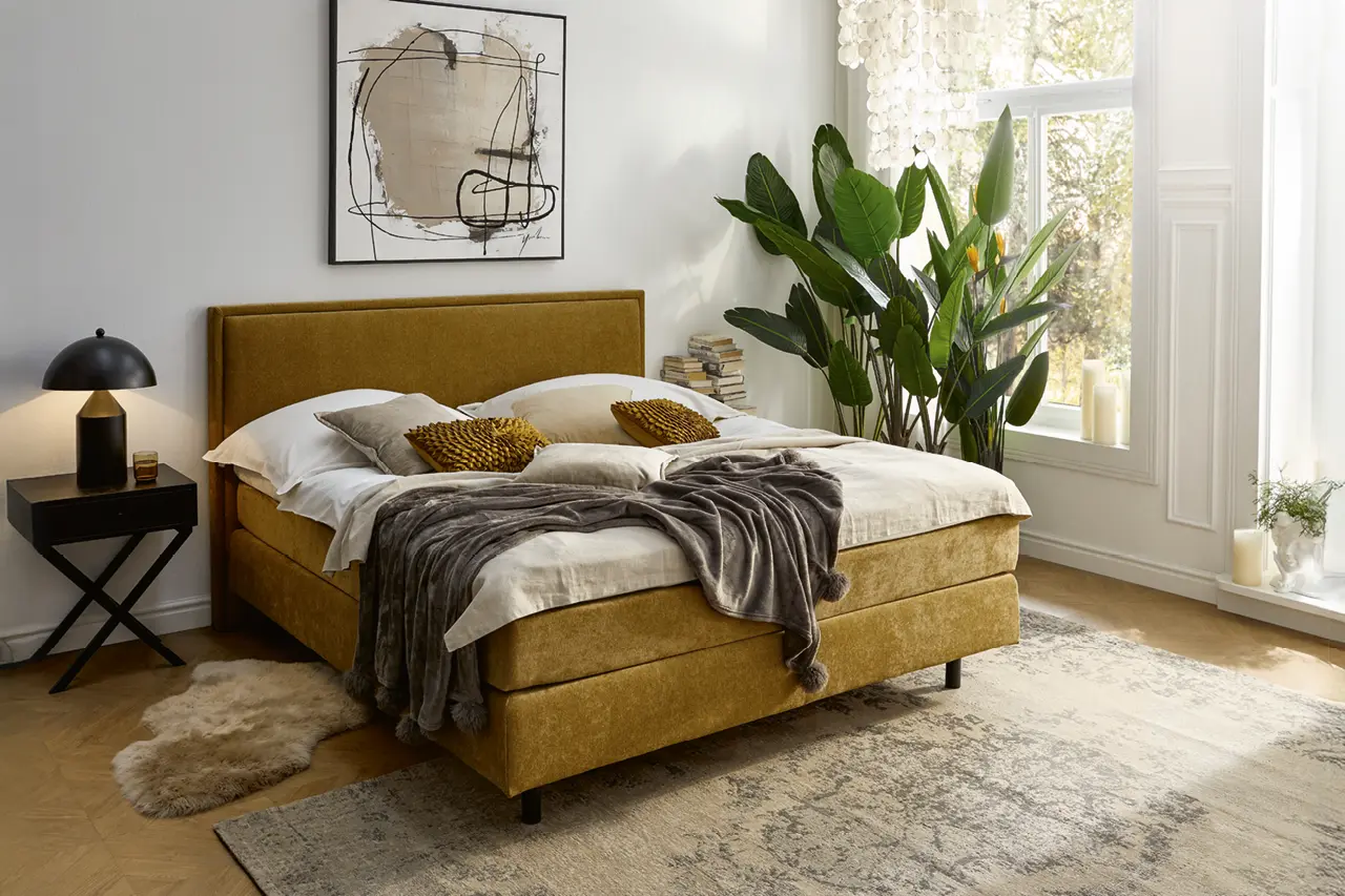 Helles Schlafzimmer mit gelbem Boxspringbett, Kissen und Decke, kombiniert mit Nachttisch und Lampe. Modernes Bett im eleganten Design, ergänzt durch Pflanzen und große Fenster für ein stilvolles Schlafambiente.